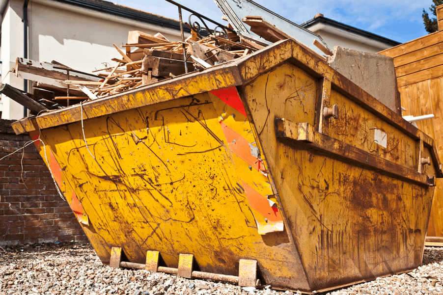 arge yellow construction dumpster filled with wood debris and scrap materials, sitting on gravel beside a house, with scratches and rust visible on its sides.
