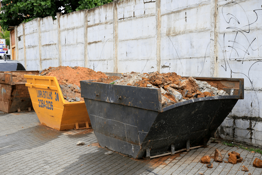 Two skips—one black and one yellow—filled with dirt, rocks, and rubble, placed on a paved surface beside a tall concrete wall with faint graffiti.