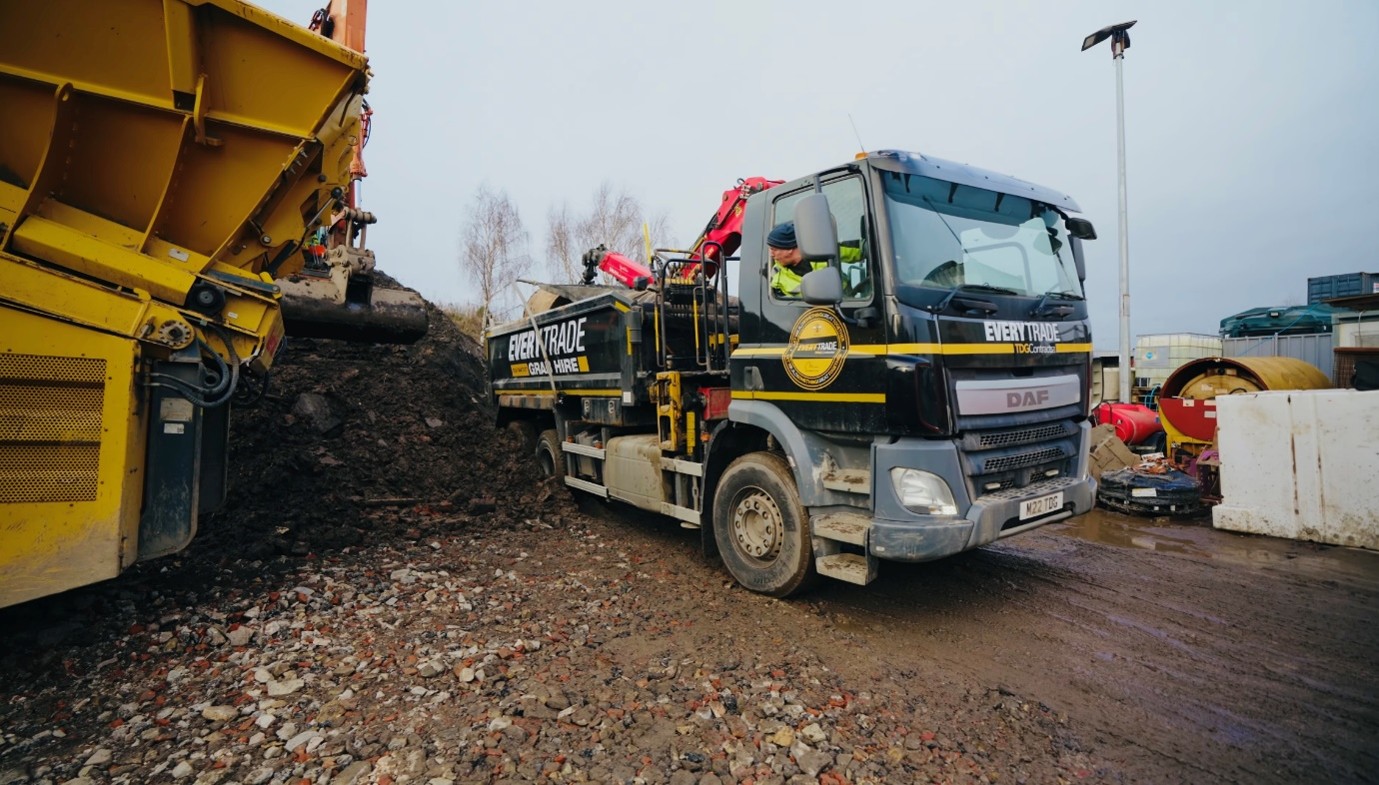 A large black DAF grab‑hire truck sits on a construction site beside a yellow soil‑screener machine. An excavator arm is actively loading soil or rubble into the truck. The ground is covered with dirt, broken bricks, and scattered industrial materials, with containers and concrete barriers visible in the background.
