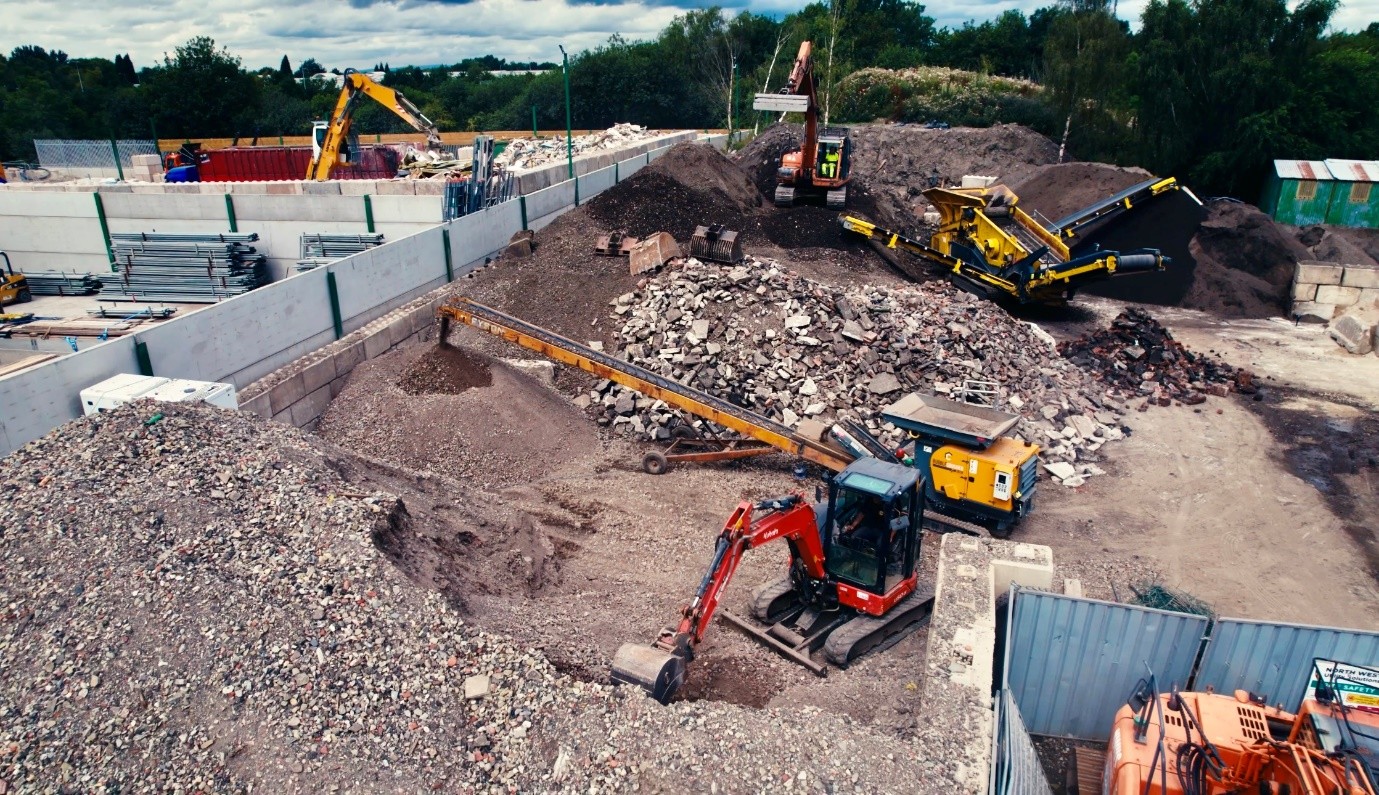Aerial view of a construction and recycling yard with multiple excavators, conveyors, and screening equipment processing large piles of crushed concrete, gravel, and aggregate materials. Surrounded by concrete storage bays and trees in the background under a cloudy sky.