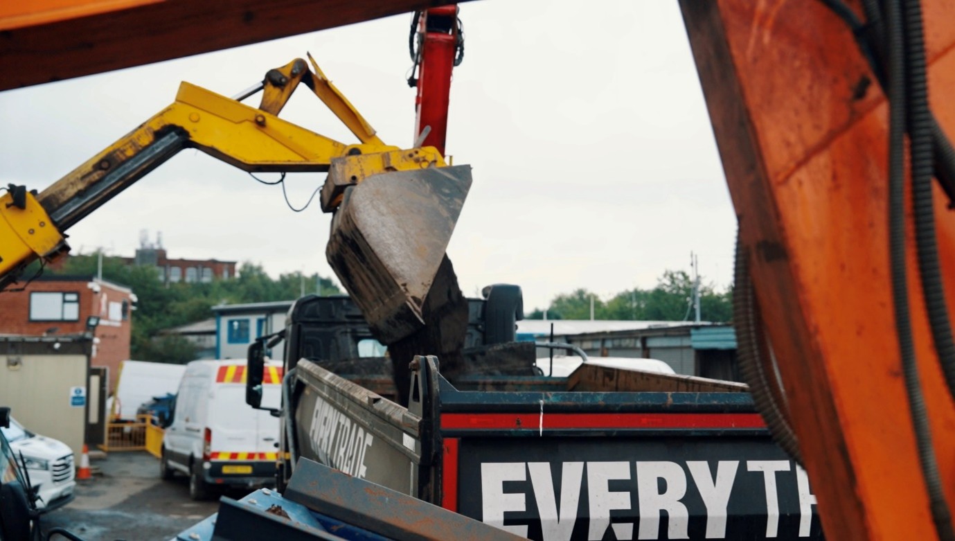 Excavator loading construction and demolition waste into a skip truck at a recycling yard. Heavy machinery is positioned above the vehicle as material is transferred into the container, with industrial buildings, parked vehicles, and equipment visible in the background.
