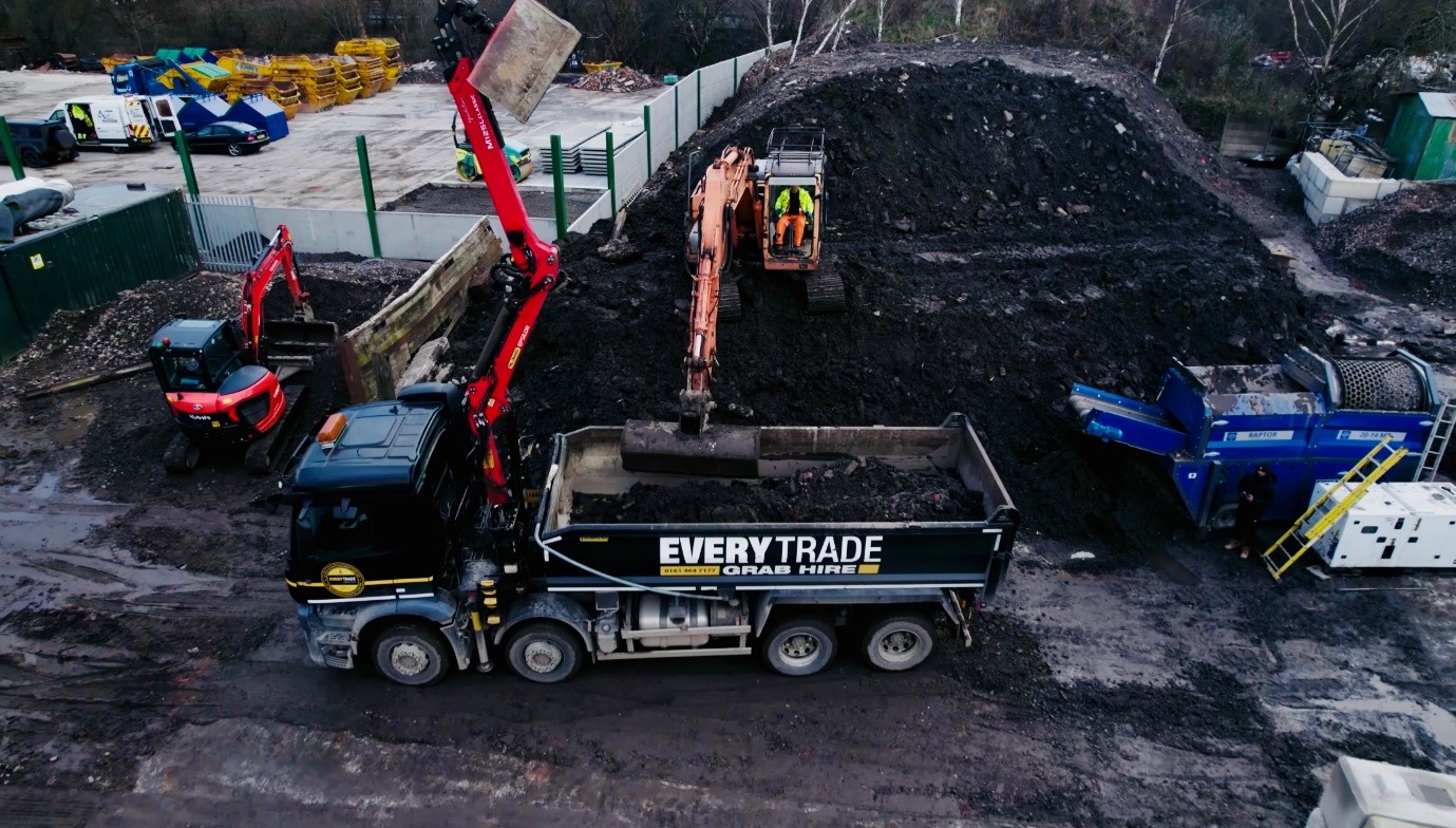 Aerial view of a recycling and aggregate processing facility, showing an Everytrade grab lorry being loaded with material by an excavator beside a large stockpile of soil and rubble, with screening machinery and additional excavators operating on site.