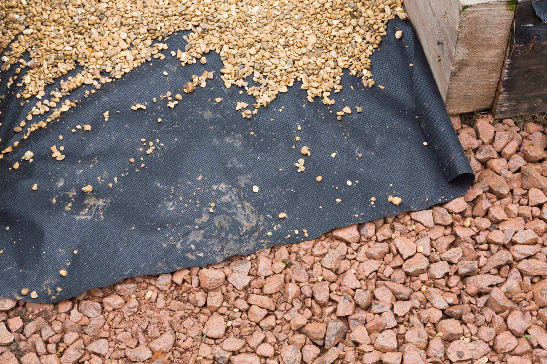 Black weed control membrane laid beneath decorative gravel, showing a layer of golden gravel above and red granite chippings below, used for weed suppression and ground stabilisation in landscaping projects.
