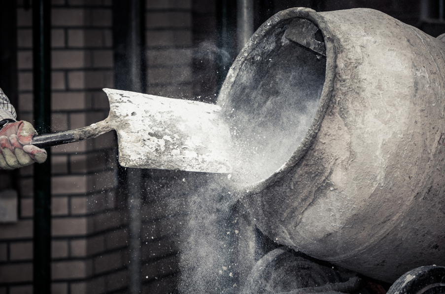 Close-up of a cement mixer drum with a shovel removing dry cement material, creating a cloud of dust during concrete preparation on a construction site