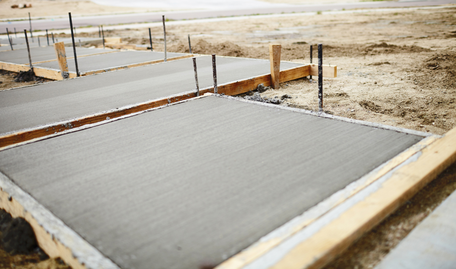 Freshly finished concrete slabs set within timber formwork on a construction site, with smooth trowelled surfaces, staking supports, and surrounding prepared ground visible in the background