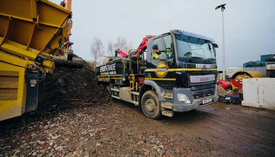 Grab lorry unloading aggregate at a recycling and waste management yard, with construction materials being tipped onto a large stockpile beside processing equipment