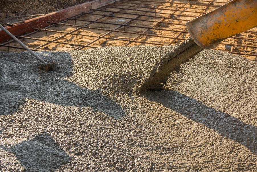 Wet concrete being poured from a chute onto a reinforced foundation slab, with steel rebar mesh visible beneath and a worker’s tool spreading the concrete across the surface